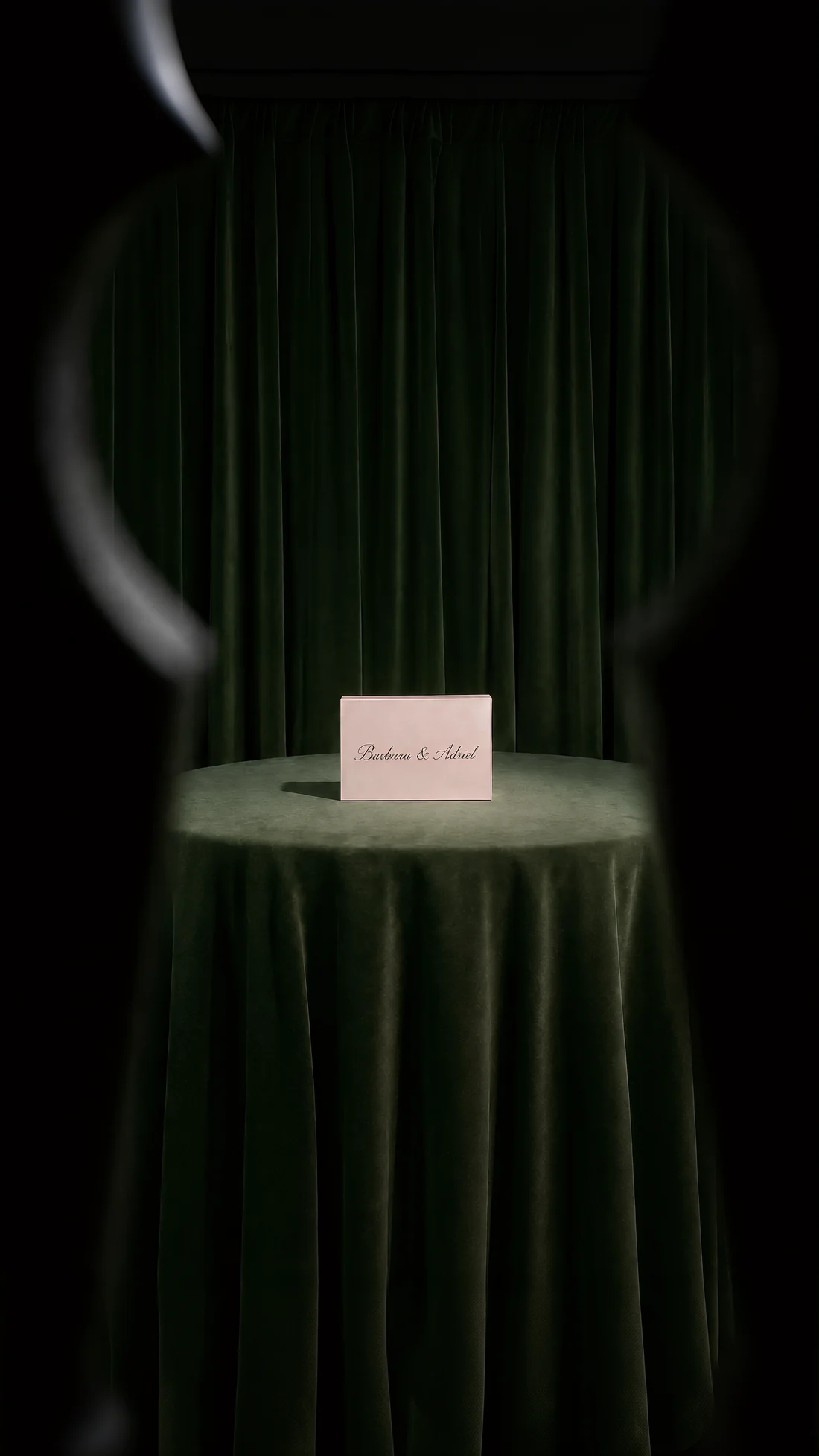 Una tarjeta con los nombres de Bárbara y Adriel, sobre una mesa vestida de terciopelo verde, vista a través de una mirilla